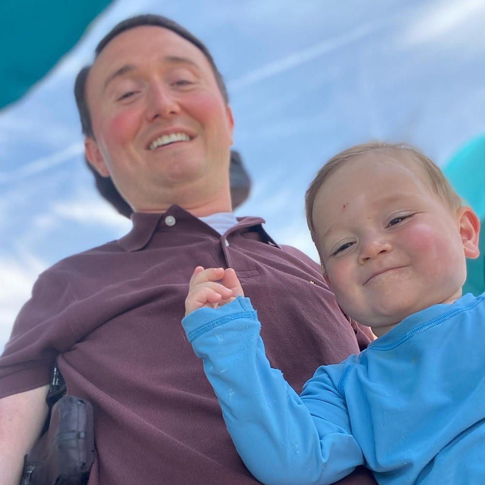 Josh and his son are outdoors backed by a blue sky streaked with white clouds. Josh has light skin, rosy cheeks, brown eyes, and short brown hair and he wears a maroon polo shirt as he sits in his black power chair. His son on his lap has light skin, brown eyes, short sandy hear, and matching rosy cheeks.