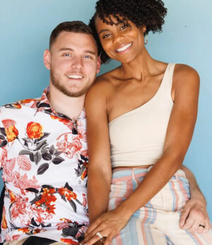 Cole and Charisma Sydnor are pictured in a photo from Forbes. Cole has light skin, short hair, a short brown beard, and a tropical short and is seated in his wheelchair. Charisma has brown skin, curly black hair, and a wedding ring visible on her left ring finger.