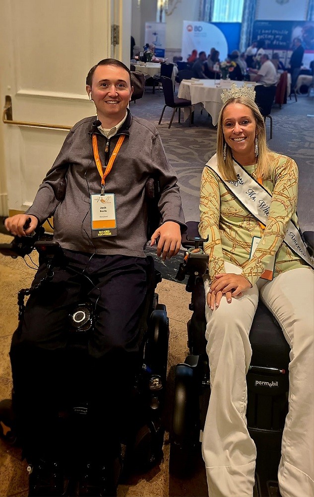 Josh Basile is at a Live to Roll conference event with Ali Ingersoll, who wears a tiara and a Ms Wheelchair America ribbon across her chest. Both are seated in their black power chairs. Josh has light skin, brown hair, and brown eyes. Ali has light skin, dark eyes, and straight shoulder-length blonde hair.