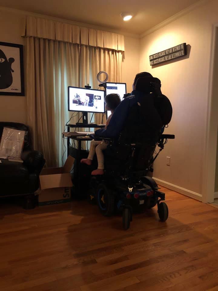 Pictured from behind, Josh Basile works at his desk with two monitors and a ringlight while seated in his black power chair on a wooden floor. His daughter sits on his lap.