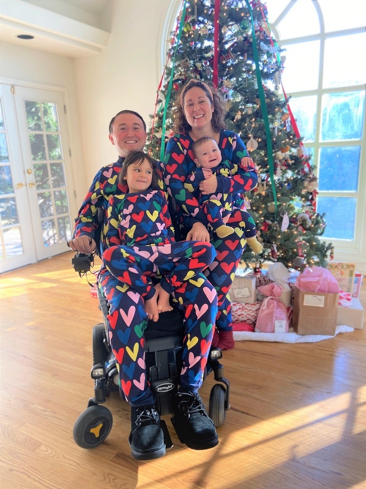 In a holiday photo, Josh, his wife, and two young children wear matching holiday pajamas with multicolored hearts on navy blue. All are smiling wide with a tall Christmas tree behind them with presents underneath it. Josh is seated in his black power chair.