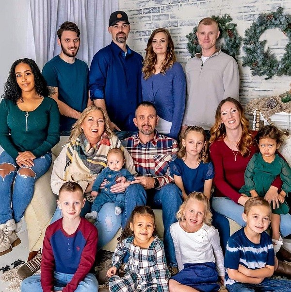 Beth Hollopeter is surrounded by standing and seated family members in shades of teal, blue, white, and red in this family portrait. Beth has light skin, blonde wavy hair to her shoulders, and red lipstick. 14 surrounding family members range in skin color, eye color, and age from baby to adult.