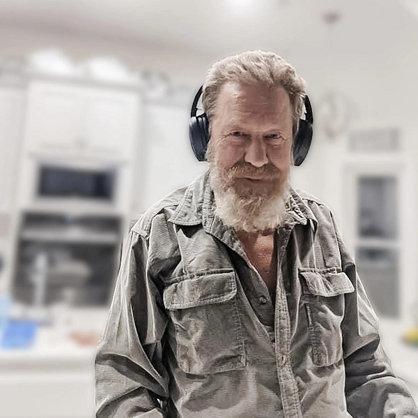 Darwin Hedges has light skin, light eyes, a sandy red and white beard, and short sandy hair. He wears a pair of large black headphones and a corduroy slate grate button-up shirt with the first two buttons undone.