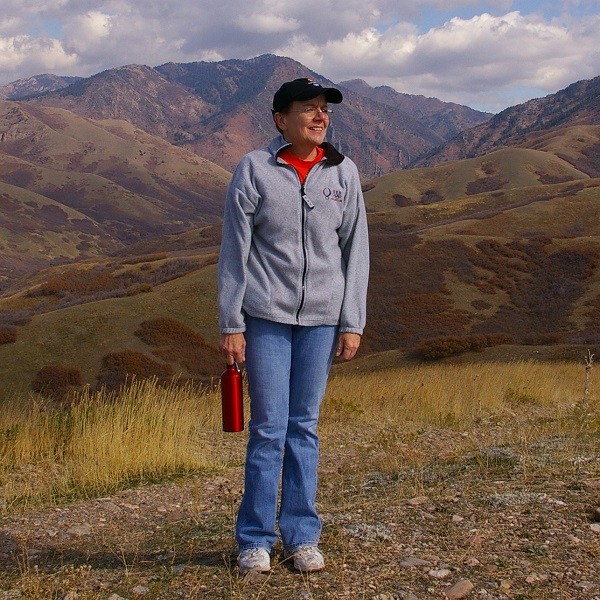 Kathleen Hartman is a slim light-skinned woman standing on a rocky trail with a beautiful mountain and valley view behind her. With glasses, a ball cap, and a red drinking thermos, Kathleen wears a fleece gray sweater, blue jeans, and hiking shoes.