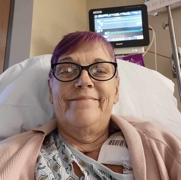 Post-transplant, Laurie Gerow takes a hospital bed selfie wearing a gown, a rose-colored sweater, black-rimmed glasses, and light fuschia-dyed hair. She has light skin and a smile.