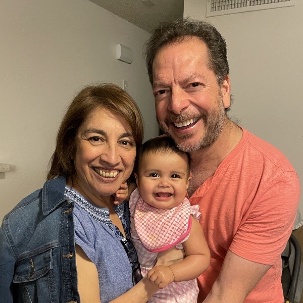 Ligia Sanchez is pictured with a man and a toddler, all grinning. Ligia has light brown skin, dark eyes, and sandy straight brown hair to her shoulders. The man has dark eyes, light skin, and a gray-brown light beard with short dark hair. The toddler has light brown skin, dark eyes, and dark short hair and wears pink gingham and a bib.