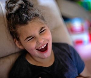 In a photo provided by the Concord Monitor, 8-year-old Lila Coe is smiling with an open mouth. She as light skin, dark hair pulled up into a bun, and a navy t-shirt.