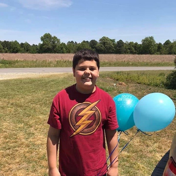 Lucas Barbour is a young teen with light skin, dark eyes, short dark hair, and a red Flash superhero logo t-shirt. He holds two blue Happy Birthday balloons in this photo as he stands outside with mowed dry grass, an empty road, and a field across from him.