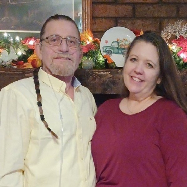 Ray Kelly is pictured in front of a fireplace mantle decorated with lights, flowers, and autumn touches. He has light skin, a short red-gray goatee, blue eyes, glasses, and a thin ponytail all the way down to his mid-chest that is tied with multiple bands to form a neat style. His female partner has light skin, light eyes, and straight brown hair and wears a magenta pink sweater.