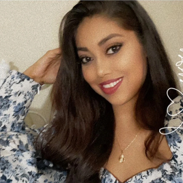 Suzel is a young woman with light brown skin, dark eyes, eyeliner, dark pink lipstick, and dark brown hair to her shoulders. She wears a flower print top and a gold locket-style necklace.