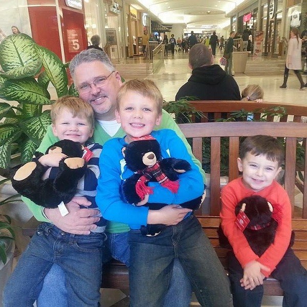 Timothy Russell is seated on a public bench inside a mall with three young boys, all smiling and clutching matching black bear teddie bears. Timothy has light skin, a light gray goatee, glasses, and short gray hair and wears jeans and a mint green sweater.