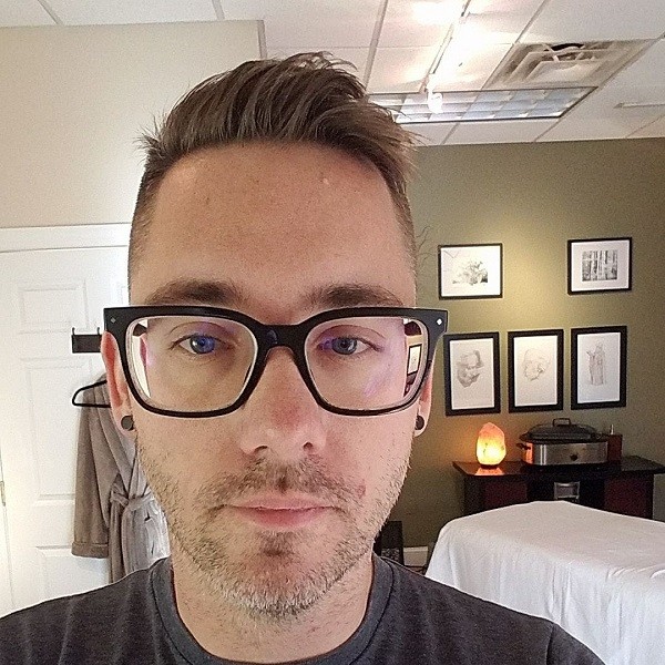 Andrew is a young man with light skin, blue-gray eyes, black-rimmed glasses, a black t-shirt, and black gauge-style earring studs. He has a short brown and gray beard. He is pictured in a massage room with a white sheet-draped table, sketch artwork on the walls, a salt lamp, and a fluffy gray-brown robe.