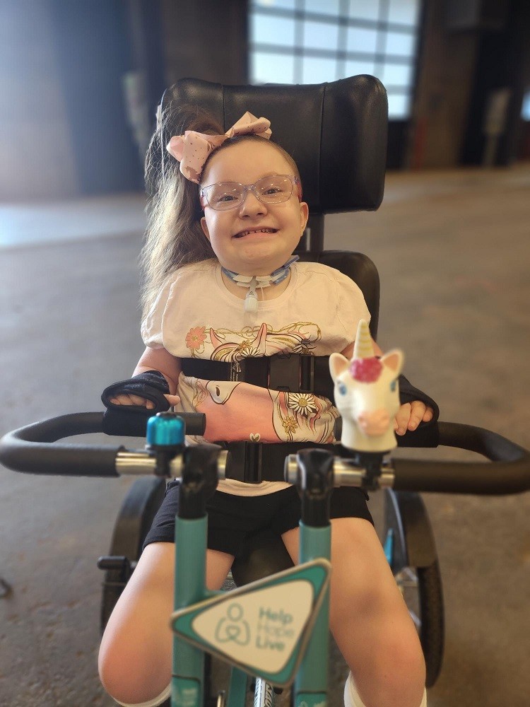 8-year-old Brooklyn has light skin, glasses, and a ponytail of wavy light brown hair with a pink bow. She is seated in her Freedom Concepts adaptive bike with the Help Hope Live logo on the front along with teal accents. She wears a star-themed necklace, a printed white t-shirt, and black shorts.