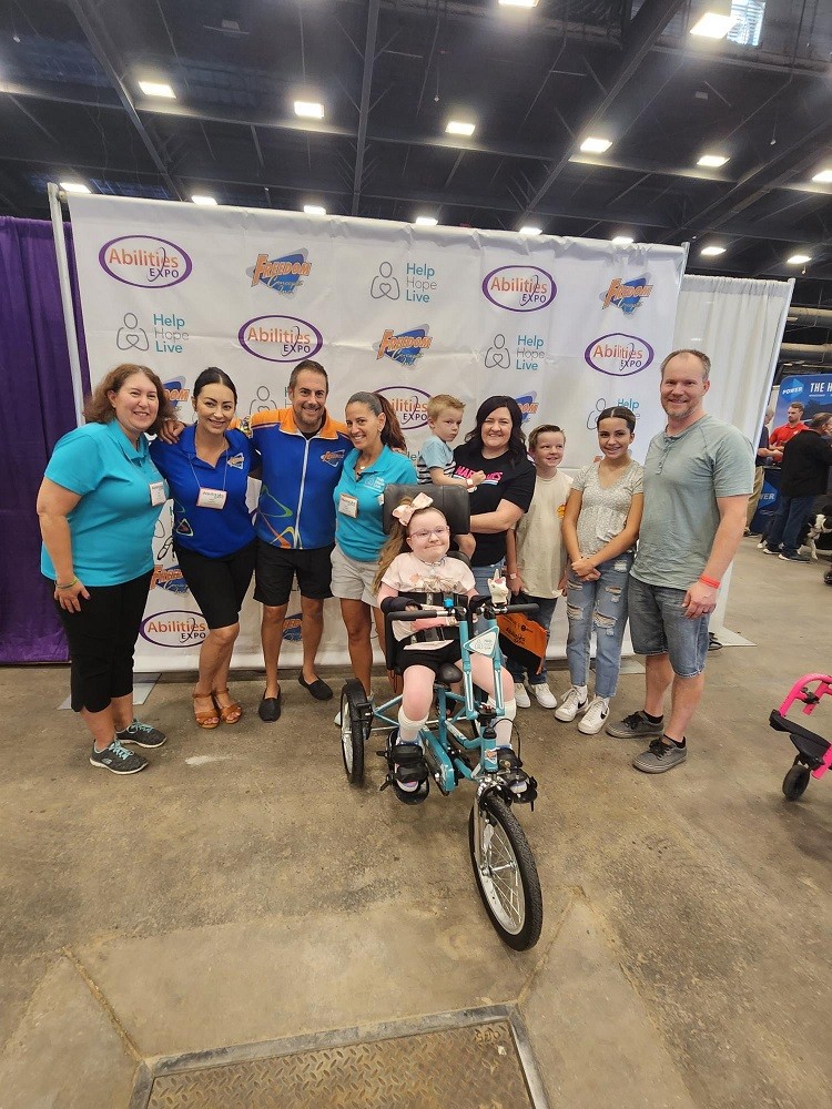 At Abilities Expo Phoenix on the expo floor, in front of a step-and-repeat banner with Freedom Concepts, Abilities Expo, and Help Hope Live logos, Help Hope Live staff members in bright teal polo shirts pose with two Freedom Concepts staff members in bright blue polo shirts along with Brooklyn on her adaptive bike plus her mom, dad, and three other children.