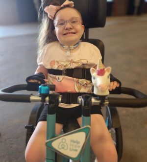 8-year-old Brooklyn has light skin, glasses, and a ponytail of wavy light brown hair with a pink bow. She is seated in her Freedom Concepts adaptive bike with the Help Hope Live logo on the front along with teal accents. She wears a star-themed necklace, a printed white t-shirt, and black shorts.