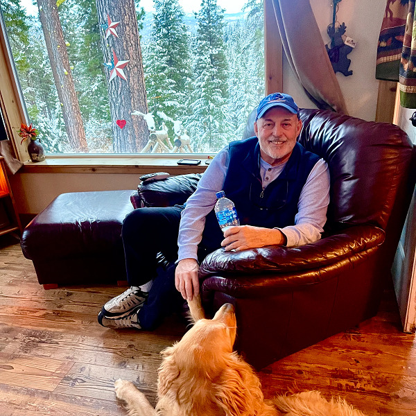 Charlie has light skin, a short white-gray beard, dark eyes, and a blue baseball cap. He wears a violet long-sleeved shirt and a blue vest. He is seated in a red leather armchair with a golden retriever lying near him. The room he is in has wooden floors and a big window with a view of a beautiful vista of snow-capped pine trees.