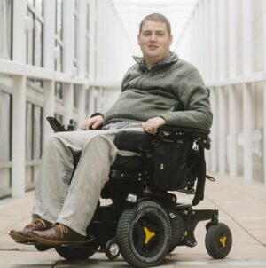 In a photo from Andrew Spear for Scientific American Mind, Ian Burkhart is seated in his black power chair in a windowed hallway. He has light skin, short brown hair, blue eyes, a button-up navy and white plaid shirt under a muted forest green fleece, khaki pants, and brown shoes.