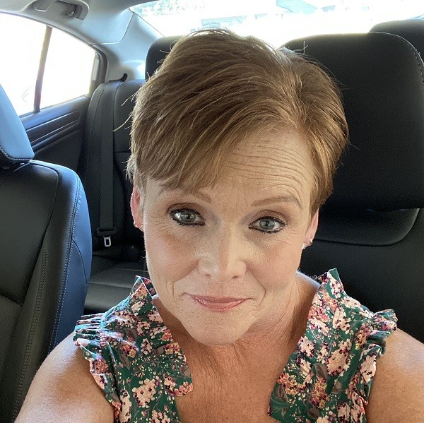 Lois has light skin, blue eyes, mascara, and peach lipstick. She has short sandy brown hair with earrings and a sleeveless floral blouse. She is taking a selfie in the driver seat of a car.