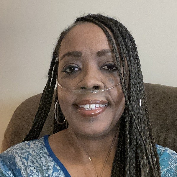 Sharnet has brown skin, brown eyes, glossy mauve lips, and long braided black hair with some gray. She wears hoop earrings and a blue patterned blouse. She has an oxygen tube connected to her nose.