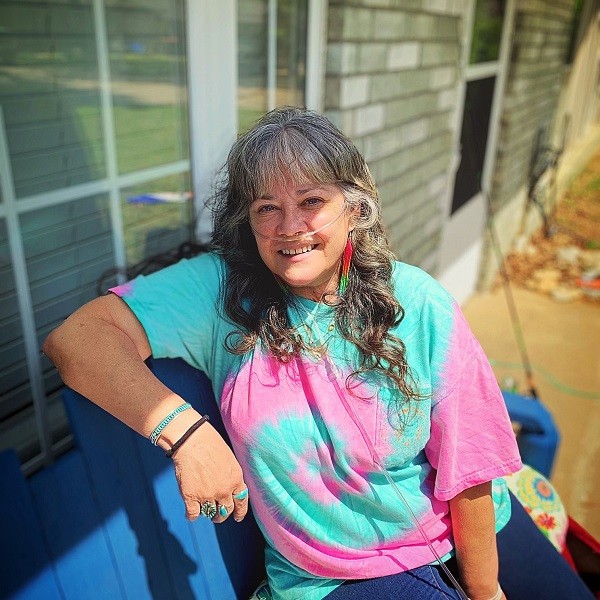 Stacia has light skin, light eyes, and curly gray hair to her chest with bangs. She wears a bright turquoise and pink tie dye t-shirt, turquoise and silver rings, and long beaded earrings. She has an oxygen tube connected to her nose. She is seated on an outdoor porch half illuminated by sunlight.