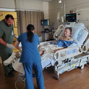 Post-transplant, Steven lies in a hospital bed with a blue hospital gown and multiple wires connected to his chest and neck. Two nurses in scrubs and gloves maneuver equipment near his feet. Steven has light skin and short gray hair.