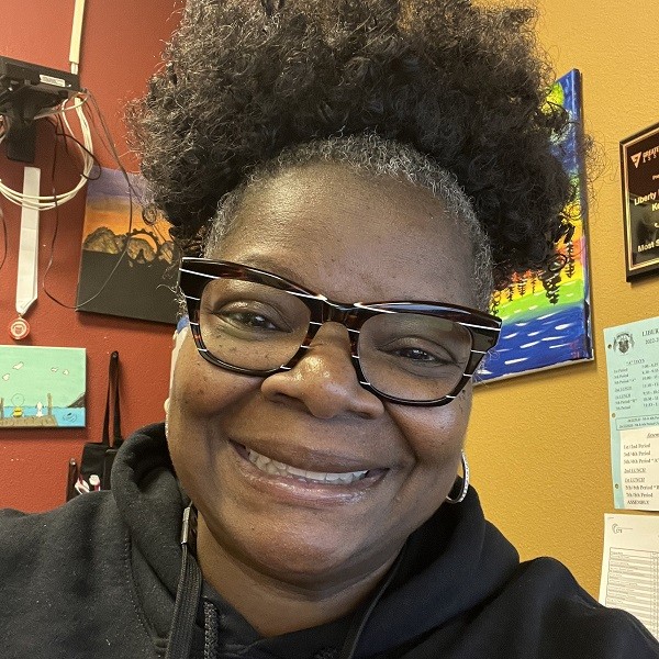 Tanisha has brown skin, brown eyes, and a big smile. She has white-striped vintage-shaped dark brown glasses and hoop earrings and wears a black hoodie. Her hair is short and curly, brown threaded with silver.