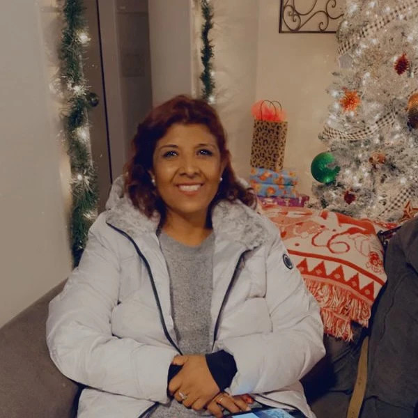 Liver transplant recipient Veronica is seated on a couch with holiday lights and a silver Christmas tree visible behind her. She has light brown skin, dark eyes, shoulder length red-brown hair, a gray t-shirt, and a warm white quilted coat.