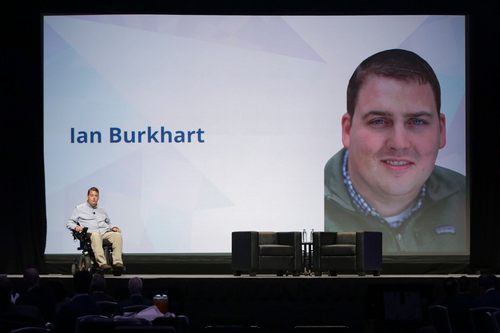 Ian Burkhart is on a stage giving a presentation with his name on a large slide behind him along with a close-up portrait. Ian has light skin, short brown hair, and blue eyes. On stage, he is seated in his black power chair with a light blue collared shirt, khaki pants, and brown shoes. There are two empty black leather chairs and two bottles of water on a black table on the other end of the stage.