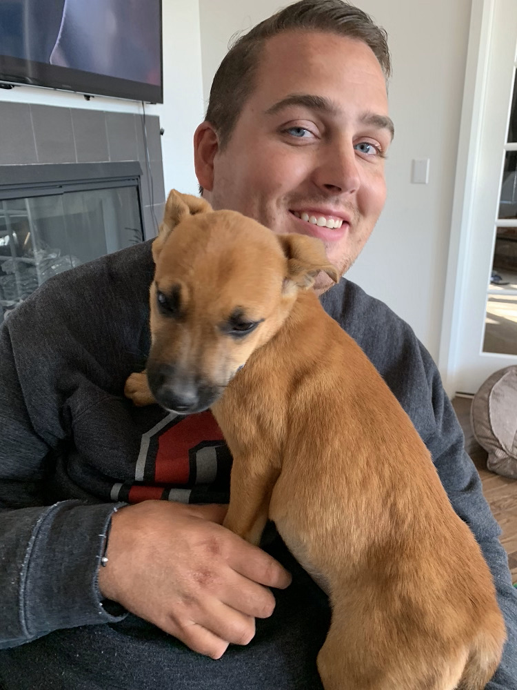 Ian Burkhart has light skin, blue eyes, and short brown hair. He is inside a home seated in his power chair, not pictured, with a small brown dog on his lap.