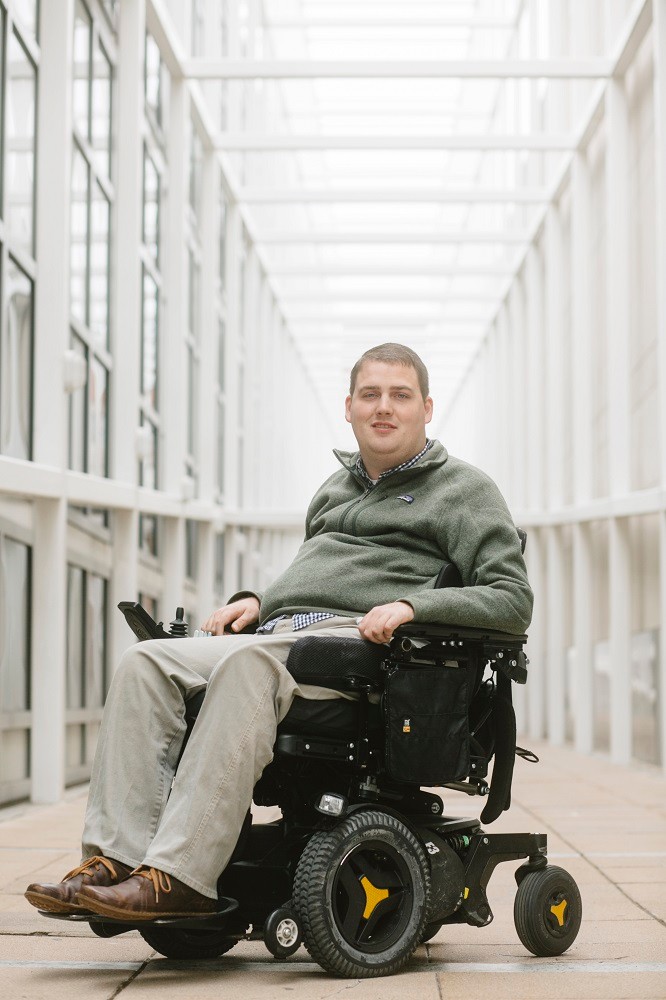 In a photo from Andrew Spear for Scientific American Mind, Ian Burkhart is seated in his black power chair in a windowed hallway. He has light skin, short brown hair, blue eyes, a button-up navy and white plaid shirt under a muted forest green fleece, khaki pants, and brown shoes.