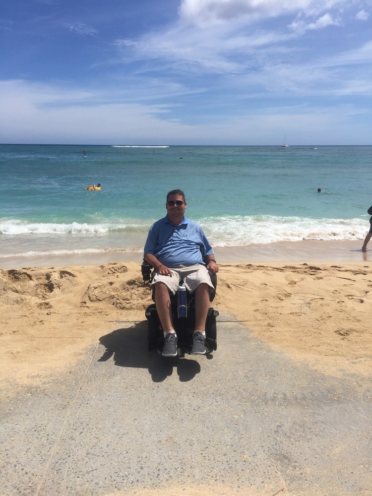 Ian Burkhart has parked his power chair on a sidewalk leading right into the sand of a beach with a light blue ocean visible behind him. He has light skin, sunglasses, short brown hair, a blue striped polo shirt, khaki shorts, and gray sneakers.