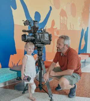 3-year-old Zeke Clark is in a building lobby with a cameraman from FOX 29 who is squatting down to match Zeke's height next to a tall black professional video recording camera. Zeke has light skin, short light blonde hair, a white sleeveless top and shorts, and bare feet with his right leg amputated just past the knee and wrapped in a bandage. He is holding a FOX 29 microphone in his right hand.