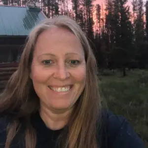 Erin Merkley takes a selfie with a cabin, forest, and beautiful pink sunset behind her. Erin has light skin, blue eyes, and sandy brown hair with some gray at the roots past her shoulders.