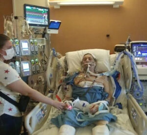 Ray Kelley is pictured in the hospital connected to a ventilator after receiving a lung transplant. He has light skin, dark hair, a hospital gown, and many medical devices and tubes connected to his body as he lies in a hospital bed. A woman reaches out to hold his hand - she has light skin and is wearing a face mask.