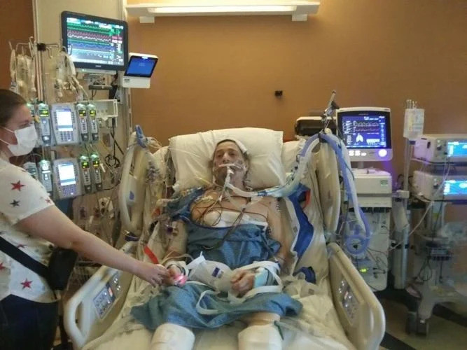 Ray Kelley is pictured in the hospital connected to a ventilator after receiving a lung transplant. He has light skin, dark hair, a hospital gown, and many medical devices and tubes connected to his body as he lies in a hospital bed. A woman reaches out to hold his hand - she has light skin and is wearing a face mask.