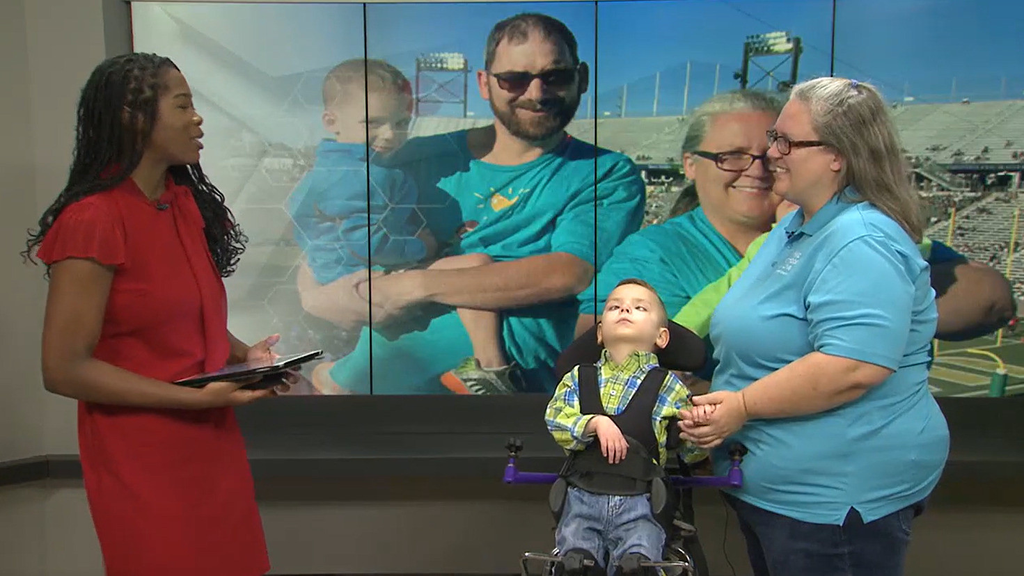 During an interview with News 4 in Dothan, Alabama, an interviewer with brown skin and a red dress speaks to Patricia King, who has light skin, gray hair, glasses, and a teal Help Hope Live shirt, as she stands next to her son, Zackariah Robinson, who is a child with light skin and short brown hair in a standing frame mobility aid. Behind them is a screen that shows Trish next to a man with light skin and short brown hair who holds Dale, Zackariah's twin brother, in his arms.