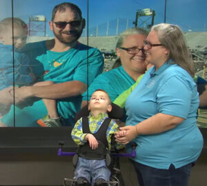 An interview with News 4 in Dothan, Alabama with Patricia King, who has light skin, gray hair, glasses, and a teal Help Hope Live shirt, as she stands next to her son, Zackariah Robinson, who is a child with light skin and short brown hair in a standing frame mobility aid. Behind them is a screen that shows Trish next to a man with light skin and short brown hair who holds Dale, Zackariah's twin brother, in his arms.