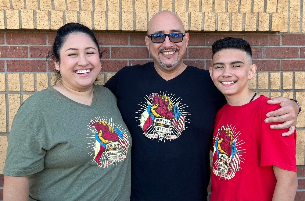 Kevin and his two children are all grinning and wearing matching t-shirts in front of a brick wall outdoors. Kevin's daughter has light brown skin, dark hair in a bun, and dark eyes. Kevin has light brown skin, a bald head, a salt-and-pepper goatee, and black-rimmed sunglasses. His son has light brown skin, dark eyes, and black hair with the sides shaved. Their t-shirts bear a logo that is a stylized hand holding a heart with the Texas and American flags and the text Heart of a Warrior Team Lopez.