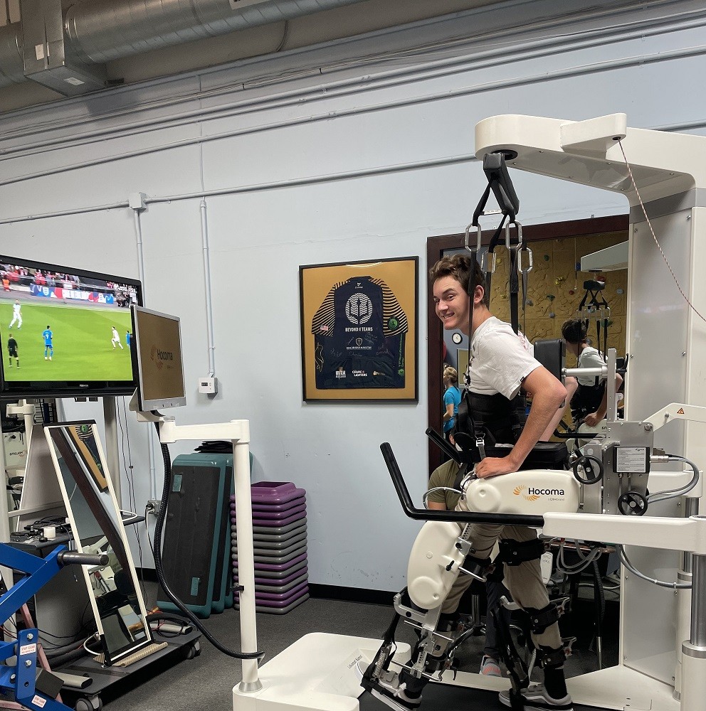 Collin Bosse uses a lokomat training device after a spinal cord injury. The device suspends him from above in a position so he can walk with assistance inside the device. Collin is a young man with light skin, short brown hair, a goofy expression, and a white t-shirt. He is watching a soccer game while he walks.