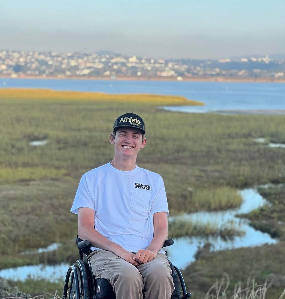 Spinal cord injury survivor Collin Bosse is a 23-year-old with light skin and a black snapback who is seated in his black wheelchair with his hands in his lap wearing cargo pants and a white t-shirt. Behind him is a marshland area.