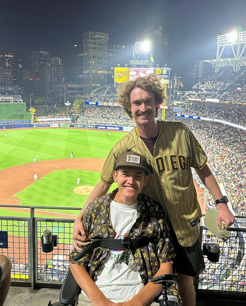 Collin Bosse is at a baseball game with a friend. He is seated in his wheelchair and strapped in across the chest and he has light skin, a gray fitted cap, a white shirt, and a printed open button-down shirt over it. His friend is standing behind him and has light skin, curly brown hair, a mustache, shorts, and a tan San Diego baseball jersey. Behind them is a baseball stadium at night with players on the diamond and a sparkling green field under the stadium lights.