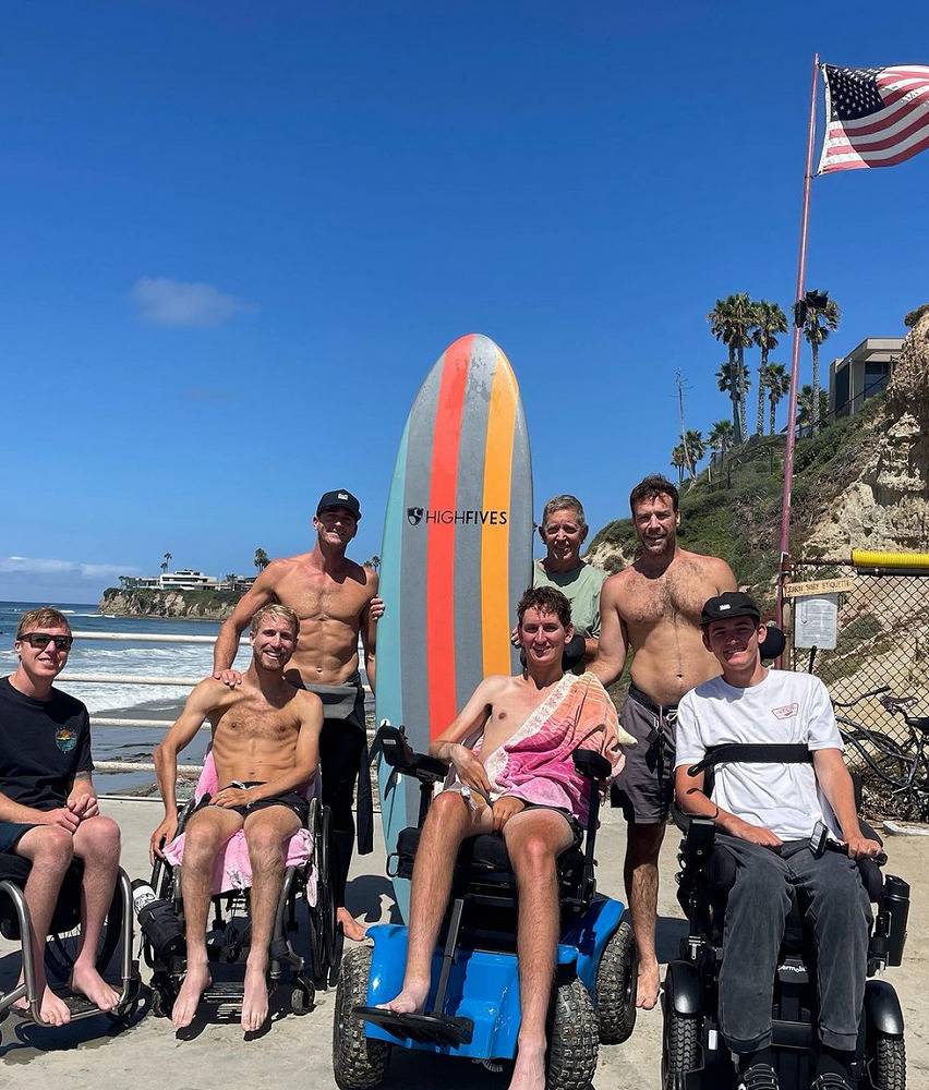 Collin Bosse is on the beach with ocean water, blue skies, and a California coastal hill visible behind him. He is with six other men close to him in age, half of whom are seated in their wheelchairs or power chairs like Collin is. One of the standing men is holding a multi-colored striped surfboard that extends above his head and reads HIGH FIVES. Collin has light skin, a black ball cap, a white t-shirt, black pants, and a black strap across his chest connecting him to his black power chair. The other men are primarily shirtless with board shorts.