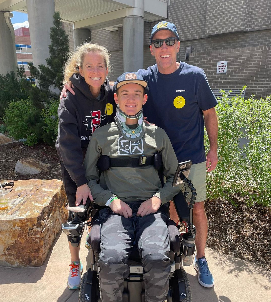 Collin and his parents are outdoors with a building and some manicured plants visible behind them in the sun. Collin is a young man with light skin, a ball cap, a gray long-sleeved shirt, and long pants who is seated in his black power chair with a strap across his chest and a neck brace. His mom has light skin and curly blonde hair and wears sneakers, an athletic hoodie, and capri-length athletic pants. His dad wears sneakers, shorts, and a navy t-shirt with a ball cap and sunglasses.