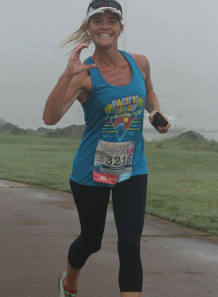 Holly Bosse, mom of client Collin Bosse, is running a half-marathon outdoors dressed in a blue tank top and black capri athletic pants with sneakers. She is giving a C sign with her left hand (C for Collin) and has light skin and blonde hair in a ponytail.
