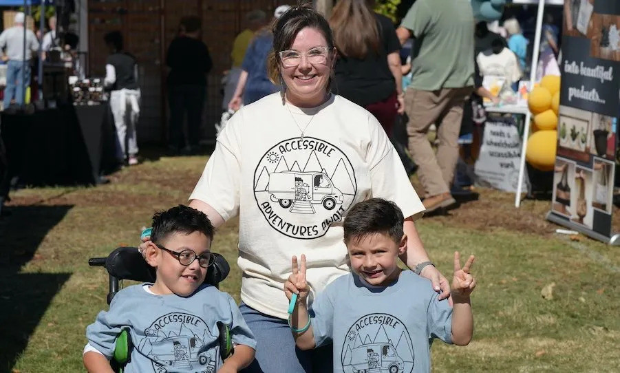 Oliver Garcia is a young kid at an outdoor festival with his mom and brother. They are wearing matching shirts that read Accessible Adventures with a logo of an accessible van and pine trees. Oliver has light brown skin, circular black glasses, short dark hair, and a black power chair. His brother is standing giving two peace signs with light brown skin and short dark hair (no glasses). Mom has light skin, dark hair pulled back, clean frame glasses, jeans, and a big smile to match Oliver's.