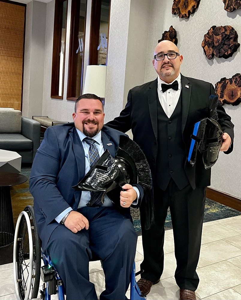 In a brightly-lit lobby, Kevin Lopez is dressed in a suit and bowtie holding an inscribed Spartan helmet in his left hand. Next to him is a man in a navy suit with a tie seated in his blue metallic wheelchair, also holding a Spartan helmet in his left hand. Kevin has light brown skin, a bald head, dark eyes, black-rimmed glasses, and a salt-and-pepper goatee. The man seated beside him has light skin, close-dropped dark hair with shaved sides, and a brown goatee.