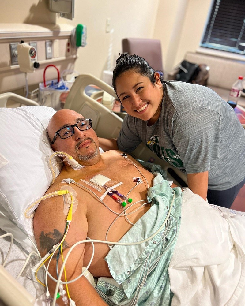 A photo of Kevin Lopez and his daughter. Kevin is lying in a hospital bed with a light green hospital gown folded down so his upper chest is exposed with many medical wires and tubes connected to it. Kevin has light brown skin, a salt-and-pepper goatee, dark eyes, black rimmed glasses, and a bald head. His daughter has light brown skin, dark hair pulled back into a bun, and dark eyes that are beautifully crinkled upward from her genuine smile.