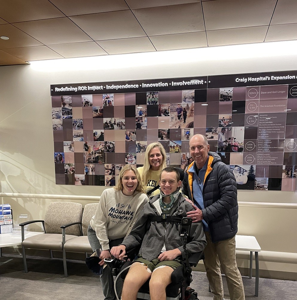 Collin and his family are at Craig Rehabilitation with a Craig infographic visible on the wall behind them. Collin is seated in his wheelchair with short dark hair, light skin, a long-sleeved gray athletic jacket, and shorts. His sister is to his right with a gray long-sleeved shirt, light skin, and blonde hair. His mom behind her is similar in appearance with light skin and blonde hair too. His dad rests his left hand on Collin's shoulder and has light skin, short gray hair, tan pants, and a black quilted cold weather jacket.