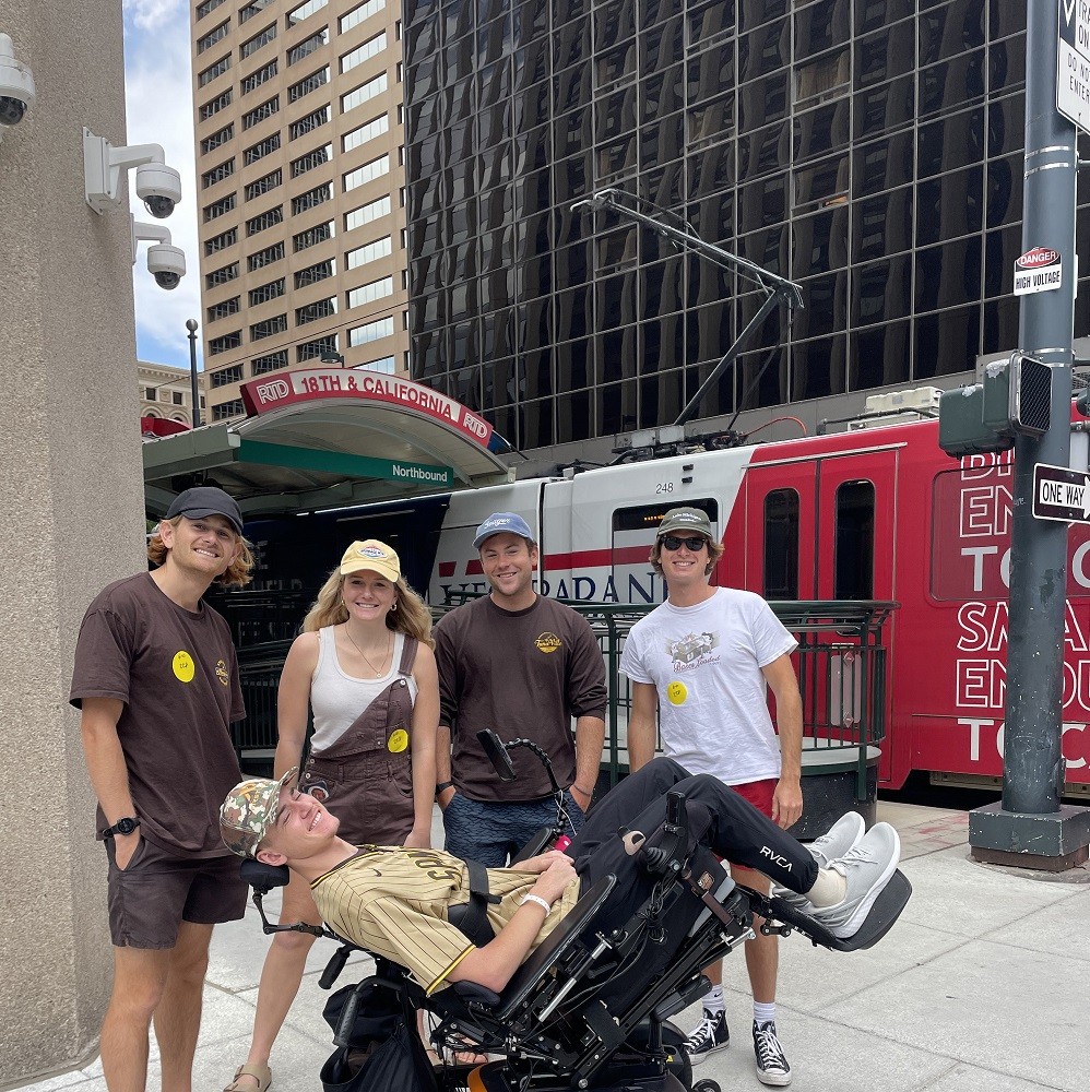 Collin Bosse is with a group of four friends in a downtown city setting with a public transportation trolley visible behind them. Collin is tilted back in his black power chair so that he is almost horizontal in his seated position. He has light skin, a camoflage ball cap, black fitted pants, a tan t-shirt, and gray sneakers. His friends are three young men with light skin and ball caps and one young woman with light skin and wavy blonde hair and a ball cap. Three of the friends wear what appears to be matching brown t-shirts with a bright yellow sticker or design on them.