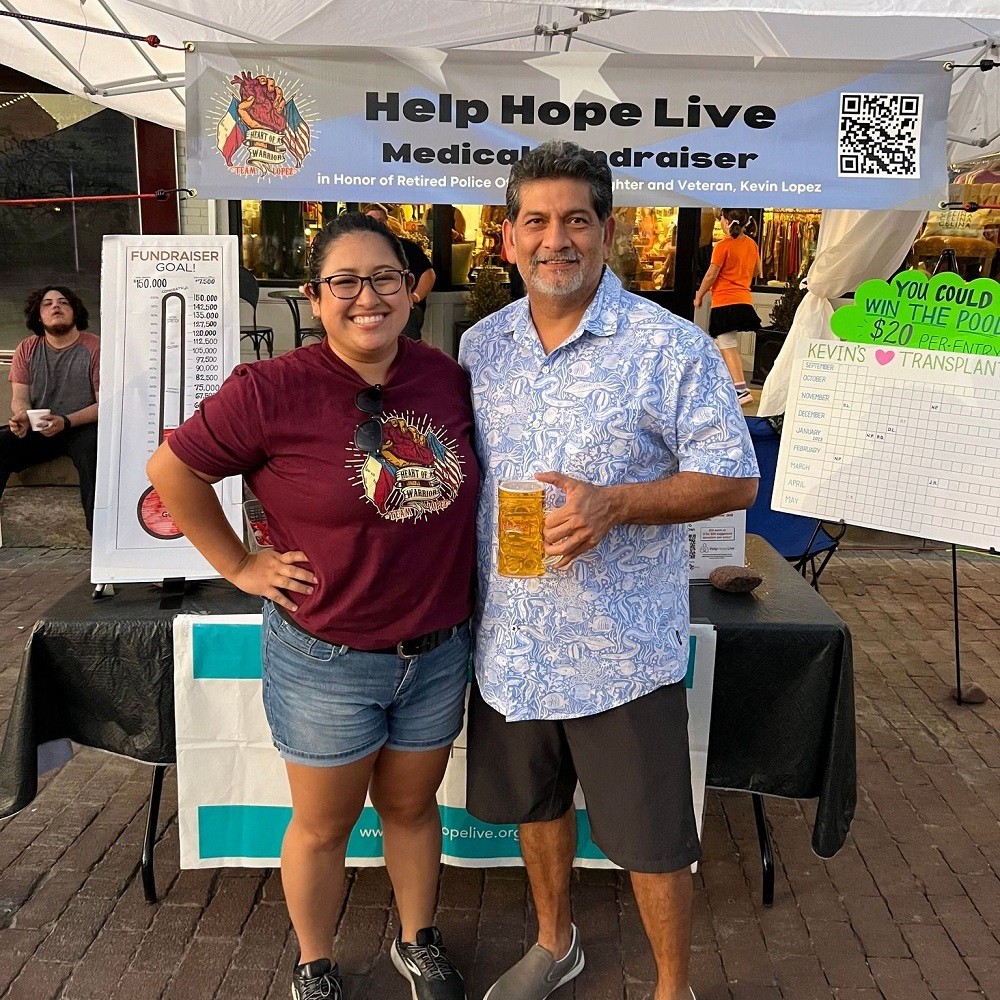 The daughter of client Kevin Lopez and a man holding a full glass of beer are standing outdoors in front of a fundraiser table with a 